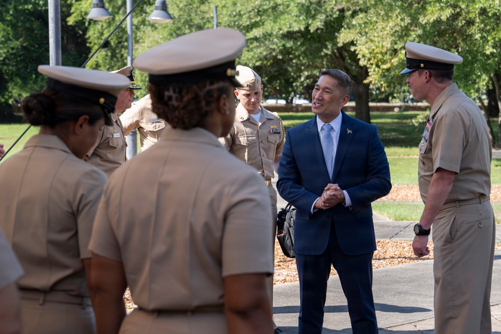 A civilian in a navy blue suit and light blue tie is engaged in a friendly conversation with several U.S. Navy personnel in tan/khaki uniforms outdoors. The setting appears to be a military installation or base, with trees and a paved walkway visible in the background. The civilian appears to be smiling and interacting warmly with the service members.