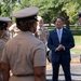 A civilian in a navy blue suit and light blue tie is engaged in a friendly conversation with several U.S. Navy personnel in tan/khaki uniforms outdoors. The setting appears to be a military installation or base, with trees and a paved walkway visible in the background. The civilian appears to be smiling and interacting warmly with the service members.