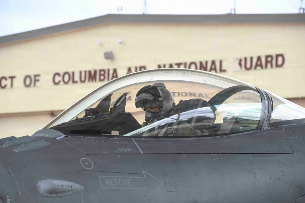 A military pilot is seated in the cockpit of a fighter jet, wearing a flight helmet and oxygen mask. The aircraft is positioned on the tarmac in front of a building bearing the sign 'District of Columbia Air National Guard.' A partial serial number '87-0243' is visible on the fuselage.