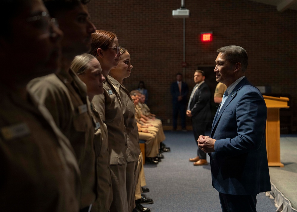 A civilian official in a blue suit is addressing or greeting a line of military personnel in tan/khaki uniforms in what appears to be an auditorium or meeting hall. The setting suggests a formal military ceremony or inspection event. Additional personnel and observers are visible seated and standing in the background.
