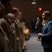 A civilian official in a blue suit is addressing or greeting a line of military personnel in tan/khaki uniforms in what appears to be an auditorium or meeting hall. The setting suggests a formal military ceremony or inspection event. Additional personnel and observers are visible seated and standing in the background.