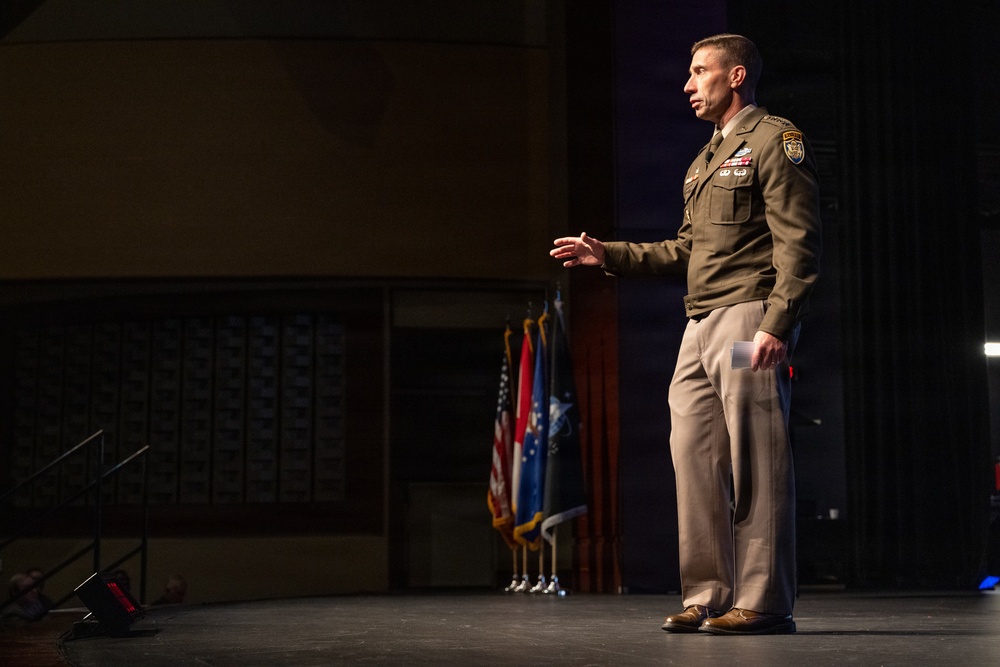 A military officer in olive green Army Service Uniform stands on a stage delivering a speech or briefing. He is gesturing with his right hand while holding papers in his left, positioned in front of a row of military and national flags. The setting appears to be a formal auditorium or theater venue.