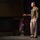 Lt. Gen. Stanton Speaks at DAFITC A military officer in olive green Army Service Uniform stands on a stage delivering a speech or briefing. He is gesturing with his right hand while holding papers in his left, positioned in front of a row of military and national flags. The setting appears to be a formal auditorium or theater venue.