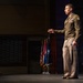 A military officer in olive green Army Service Uniform stands on a stage delivering a speech or briefing. He is gesturing with his right hand while holding papers in his left, positioned in front of a row of military and national flags. The setting appears to be a formal auditorium or theater venue.