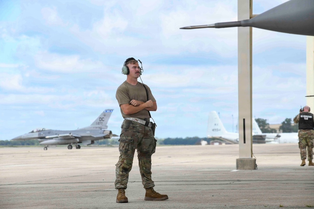 A military airman in OCP (Operational Camouflage Pattern) uniform stands on a flight line apron with arms crossed, wearing aviation headset/hearing protection. In the background, an F-16 Fighting Falcon with 'DC' tail markings (indicating DC Air National Guard) is visible on the taxiway. A second personnel member wearing a vest labeled 'IG INSPECTION TEAM' is visible near a hangar structure on the right.