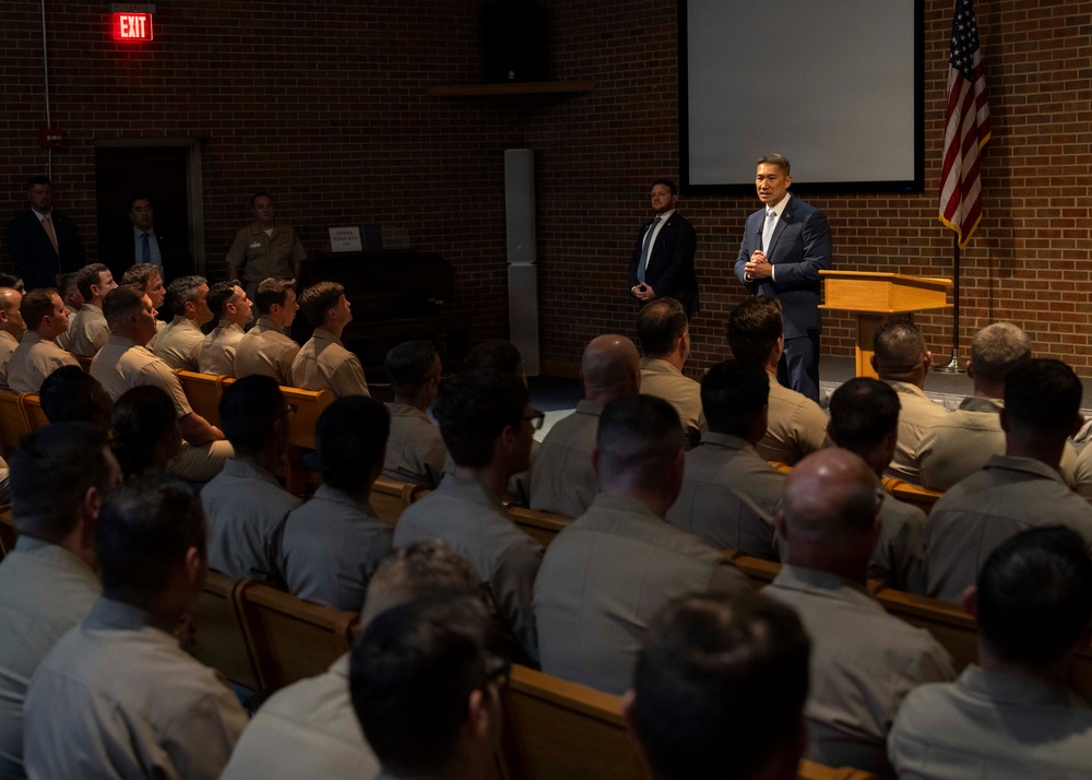 A civilian official in a dark blue suit is addressing a large group of military personnel seated in an auditorium-style room. The audience is wearing tan/khaki uniforms, suggesting a U.S. military branch briefing or lecture. An American flag and a projection screen are visible behind the speaker near a wooden podium.