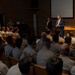 A civilian official in a dark blue suit is addressing a large group of military personnel seated in an auditorium-style room. The audience is wearing tan/khaki uniforms, suggesting a U.S. military branch briefing or lecture. An American flag and a projection screen are visible behind the speaker near a wooden podium.