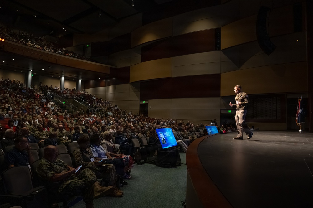 A military officer in uniform is delivering a presentation on a large auditorium stage to a packed audience of hundreds of attendees, many of whom appear to be in military uniforms. The venue is a modern, multi-level auditorium with both floor-level and balcony seating fully occupied. Presentation screens are visible near the stage displaying slides.