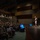 Lt. Gen. Stanton at DAFITC A military officer in uniform is delivering a presentation on a large auditorium stage to a packed audience of hundreds of attendees, many of whom appear to be in military uniforms. The venue is a modern, multi-level auditorium with both floor-level and balcony seating fully occupied. Presentation screens are visible near the stage displaying slides.