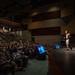 A military officer in uniform is delivering a presentation on a large auditorium stage to a packed audience of hundreds of attendees, many of whom appear to be in military uniforms. The venue is a modern, multi-level auditorium with both floor-level and balcony seating fully occupied. Presentation screens are visible near the stage displaying slides.