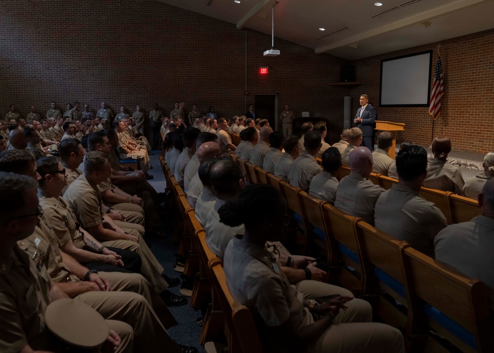 A civilian speaker in a dark suit addresses a large audience of military personnel in khaki uniforms seated in an auditorium-style room. The setting appears to be a formal briefing or lecture at a military installation, with an American flag and projection screen visible at the front. The room is filled to capacity with uniformed service members facing the podium.