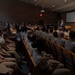 A civilian speaker in a dark suit addresses a large audience of military personnel in khaki uniforms seated in an auditorium-style room. The setting appears to be a formal briefing or lecture at a military installation, with an American flag and projection screen visible at the front. The room is filled to capacity with uniformed service members facing the podium.