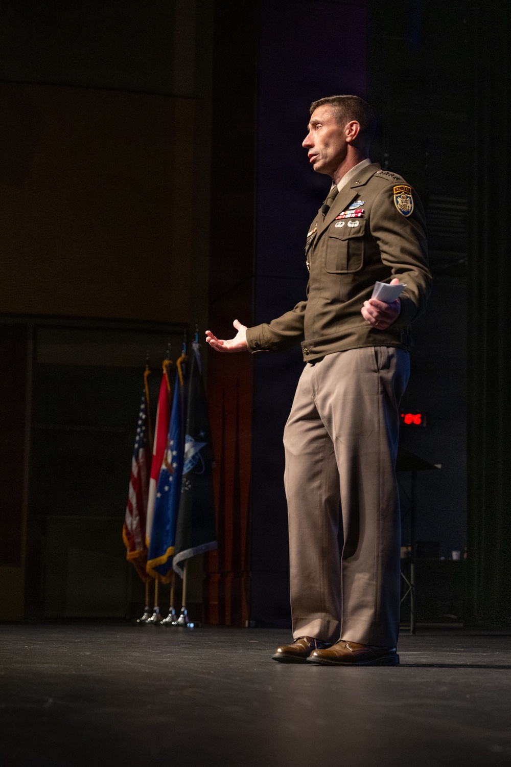 A U.S. Army officer in dress uniform stands on a stage delivering a speech or address, holding what appears to be note cards in one hand while gesturing with the other. Multiple military and U.S. flags are displayed on stands in the background. The setting appears to be a formal military ceremony or official event held in an auditorium or theater.