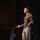 Lt. Gen. Stanton speaks at DAFITC A U.S. Army officer in dress uniform stands on a stage delivering a speech or address, holding what appears to be note cards in one hand while gesturing with the other. Multiple military and U.S. flags are displayed on stands in the background. The setting appears to be a formal military ceremony or official event held in an auditorium or theater.