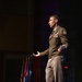 A U.S. Army officer in dress uniform stands on a stage delivering a speech or address, holding what appears to be note cards in one hand while gesturing with the other. Multiple military and U.S. flags are displayed on stands in the background. The setting appears to be a formal military ceremony or official event held in an auditorium or theater.