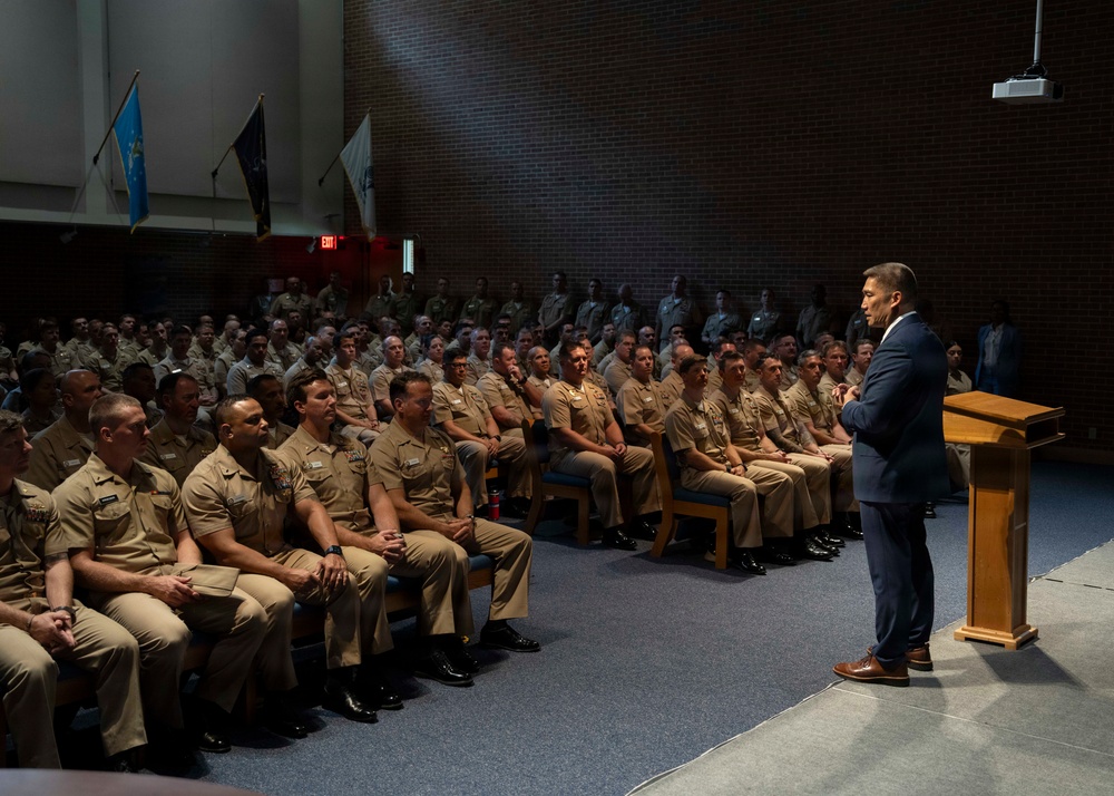 A civilian speaker in a dark suit addresses a large audience of uniformed military personnel seated in an auditorium. The personnel are wearing tan/khaki uniforms consistent with U.S. Navy service uniforms. The speaker stands at a wooden podium on a raised stage area.