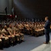A civilian speaker in a dark suit addresses a large audience of uniformed military personnel seated in an auditorium. The personnel are wearing tan/khaki uniforms consistent with U.S. Navy service uniforms. The speaker stands at a wooden podium on a raised stage area.