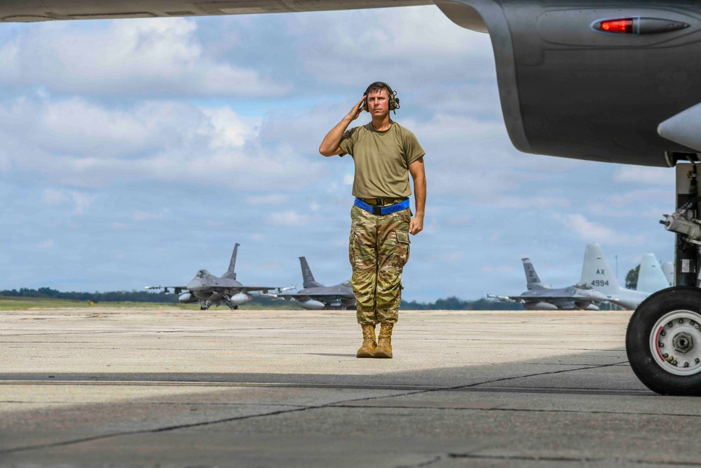 A military ground crew member wearing OCP (Operational Camouflage Pattern) uniform and hearing protection walks across a flight line tarmac, adjusting his headset. He is framed beneath the wing and landing gear of a large aircraft in the foreground. Multiple F-16 Fighting Falcon jets are visible in the background on the flight line.