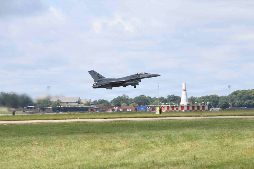 A U.S. Air Force F-16 Fighting Falcon jet is captured just after takeoff or during a low-altitude pass over a runway. The aircraft bears 'DC' tail markings and the serial number appears to read '877', indicating it belongs to the DC Air National Guard. The setting appears to be a military airfield with green grass, runway markers, and a white-and-red striped distance marker cone visible in the background.