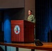 A U.S. Army officer in OCP (Operational Camouflage Pattern) uniform stands at a wooden podium on a stage, delivering a speech or briefing. The podium bears the seal of the Maneuver Center of Excellence at Fort Benning, Georgia. The setting appears to be a formal military auditorium or theater.
