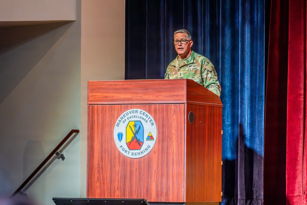 A U.S. Army officer in Army Combat Uniform (ACU/OCP pattern) is speaking at a wooden podium on a stage. The podium displays the official seal of the Maneuver Center of Excellence at Fort Benning. The setting appears to be a formal military auditorium or theater with blue and red curtains in the background.