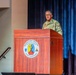 A U.S. Army officer in Army Combat Uniform (ACU/OCP pattern) is speaking at a wooden podium on a stage. The podium displays the official seal of the Maneuver Center of Excellence at Fort Benning. The setting appears to be a formal military auditorium or theater with blue and red curtains in the background.
