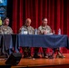 Three U.S. Army soldiers in camouflage uniforms are seated at a panel table on a stage during the 2026 Maneuver Warfighter Conference. The rightmost soldier appears to be actively speaking, gesturing with his hands, while the other two listen. The event is set against a red curtain backdrop with a conference banner visible on the left.