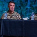 Two U.S. Army personnel are seated at a table covered with a black cloth, appearing to participate in a panel discussion or briefing. The soldier on the left, with the partial name tag reading 'SH', is speaking into a microphone, while the soldier on the right, named 'ACTON', listens attentively. Both wear OCP (Operational Camouflage Pattern) uniforms with U.S. Army tape and American flag patches.