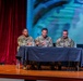 Three U.S. Army personnel in OCP (Operational Camouflage Pattern) uniforms are seated at a table on a stage, appearing to participate in a panel discussion or briefing. The setting appears to be a formal military event or town hall, held in an auditorium with a projected screen backdrop and red curtain. Name placards and microphones are visible on the table in front of the panelists.