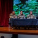 Three U.S. Army soldiers in OCP (Operational Camouflage Pattern) uniforms are seated at a table covered with a black tablecloth on a stage, appearing to participate in a panel discussion or briefing. The setting appears to be an auditorium or theater, with red curtains on the left and a large projection screen displaying a green and blue abstract image in the background. Water bottles and papers are visible on the table in front of the panelists.