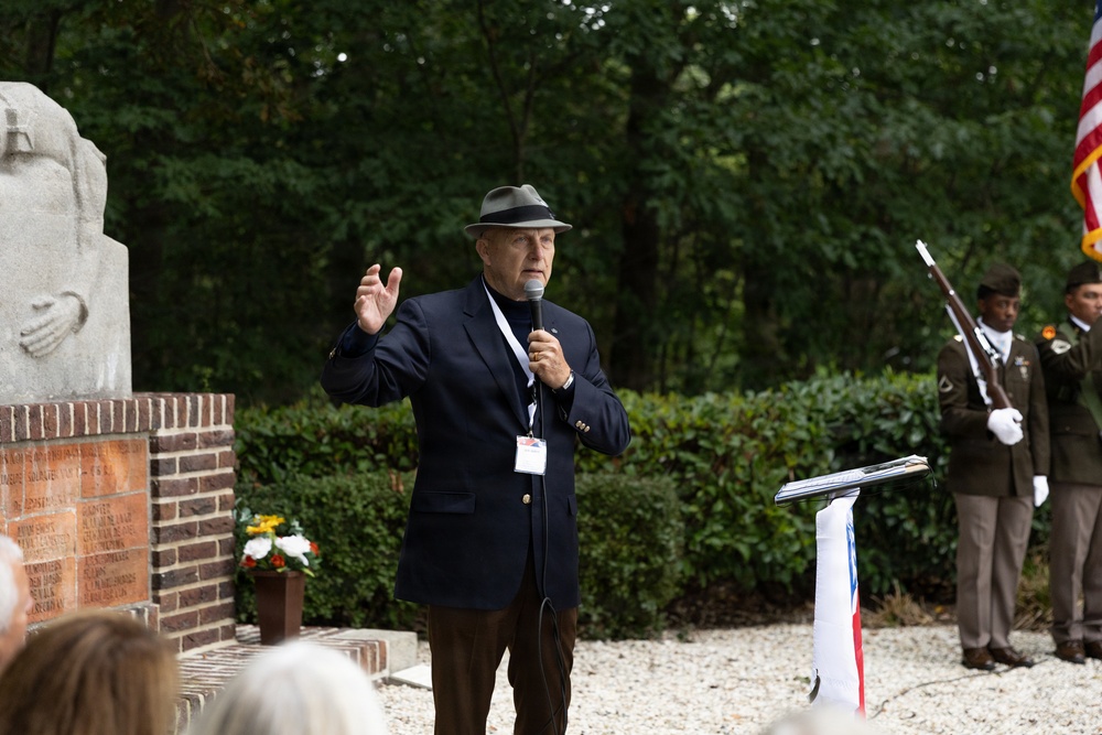 An elderly man in a navy blazer and gray fedora hat is speaking into a microphone at an outdoor memorial ceremony. Behind him stands a military honor guard soldier in olive dress uniform holding a rifle, alongside an American flag. A stone memorial monument with inscribed names and a brick base is visible to the left.