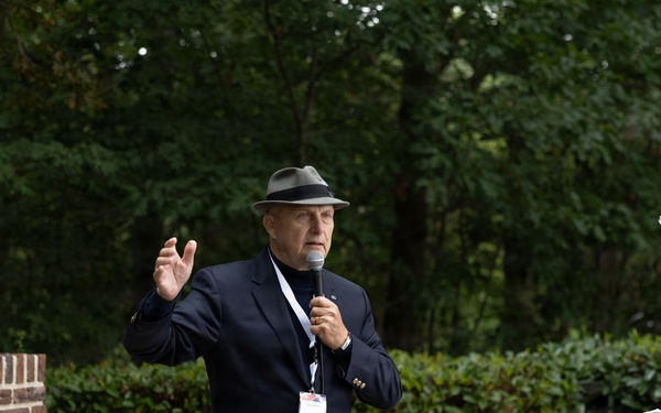 An elderly man in a navy blazer and gray fedora hat is speaking into a microphone at an outdoor memorial ceremony. Behind him stands a military honor guard soldier in olive dress uniform holding a rifle, alongside an American flag. A stone memorial monument with inscribed names and a brick base is visible to the left.