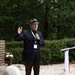 An elderly man in a navy blazer and gray fedora hat is speaking into a microphone at an outdoor memorial ceremony. Behind him stands a military honor guard soldier in olive dress uniform holding a rifle, alongside an American flag. A stone memorial monument with inscribed names and a brick base is visible to the left.