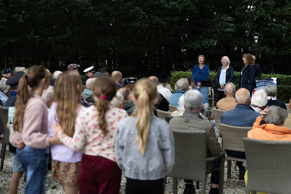 An outdoor gathering or ceremony is taking place in a garden or park setting, with a crowd of mixed ages seated and standing while three women appear to be performing or speaking at the front. A partial banner reading 'AIRBORNE' is visible on the right side, suggesting this may be a commemorative event related to WWII Airborne forces. The audience includes elderly individuals, adults, and children, consistent with a memorial or remembrance ceremony.