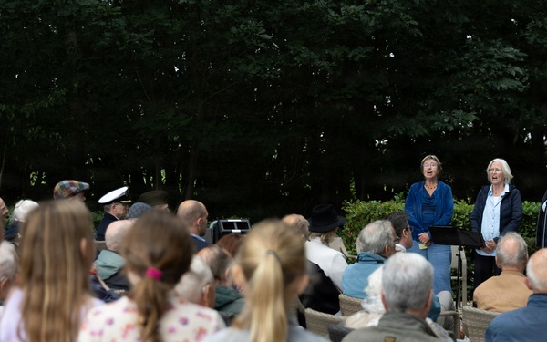 An outdoor gathering or ceremony is taking place in a garden or park setting, with a crowd of mixed ages seated and standing while three women appear to be performing or speaking at the front. A partial banner reading 'AIRBORNE' is visible on the right side, suggesting this may be a commemorative event related to WWII Airborne forces. The audience includes elderly individuals, adults, and children, consistent with a memorial or remembrance ceremony.