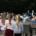 An outdoor gathering or ceremony is taking place in a garden or park setting, with a crowd of mixed ages seated and standing while three women appear to be performing or speaking at the front. A partial banner reading 'AIRBORNE' is visible on the right side, suggesting this may be a commemorative event related to WWII Airborne forces. The audience includes elderly individuals, adults, and children, consistent with a memorial or remembrance ceremony.