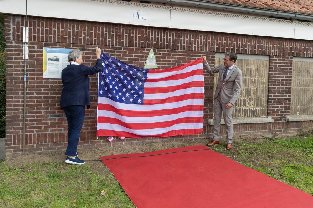 Two individuals are holding up a large American flag in front of a brick building, appearing to unveil a commemorative plaque mounted on the wall behind them. A red carpet leads toward the building, suggesting a formal ceremony is taking place. Small American flags are planted in the ground along the base of the building.