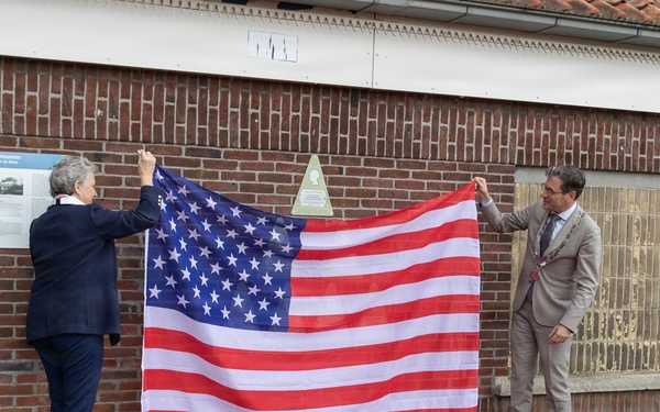 Two individuals are holding up a large American flag in front of a brick building, appearing to unveil a commemorative plaque mounted on the wall behind them. A red carpet leads toward the building, suggesting a formal ceremony is taking place. Small American flags are planted in the ground along the base of the building.