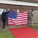 Two individuals are holding up a large American flag in front of a brick building, appearing to unveil a commemorative plaque mounted on the wall behind them. A red carpet leads toward the building, suggesting a formal ceremony is taking place. Small American flags are planted in the ground along the base of the building.
