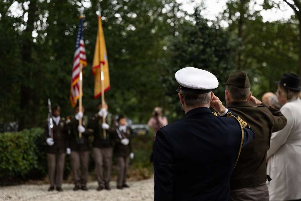 A military ceremony is taking place outdoors, with officers in the foreground rendering a salute toward a color guard in the background. The color guard is presenting both the United States flag and what appears to be a yellow/gold military or unit flag. The setting appears to be a wooded or garden area, suggesting a memorial or commemorative event.