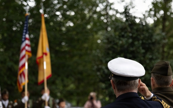 A military ceremony is taking place outdoors, with officers in the foreground rendering a salute toward a color guard in the background. The color guard is presenting both the United States flag and what appears to be a yellow/gold military or unit flag. The setting appears to be a wooded or garden area, suggesting a memorial or commemorative event.