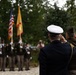 A military ceremony is taking place outdoors, with officers in the foreground rendering a salute toward a color guard in the background. The color guard is presenting both the United States flag and what appears to be a yellow/gold military or unit flag. The setting appears to be a wooded or garden area, suggesting a memorial or commemorative event.
