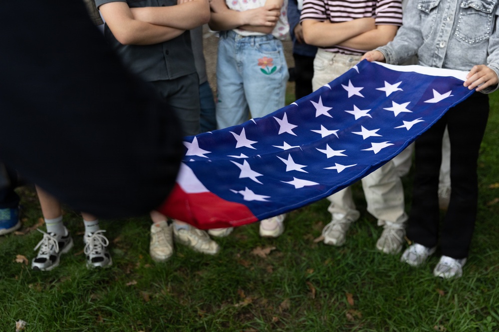 A group of people standing on grass are holding an American flag, visible from the waist down. The flag is being held horizontally, displaying the blue canton with white stars prominently. The setting appears to be an outdoor gathering or event.