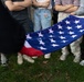 A group of people standing on grass are holding an American flag, visible from the waist down. The flag is being held horizontally, displaying the blue canton with white stars prominently. The setting appears to be an outdoor gathering or event.