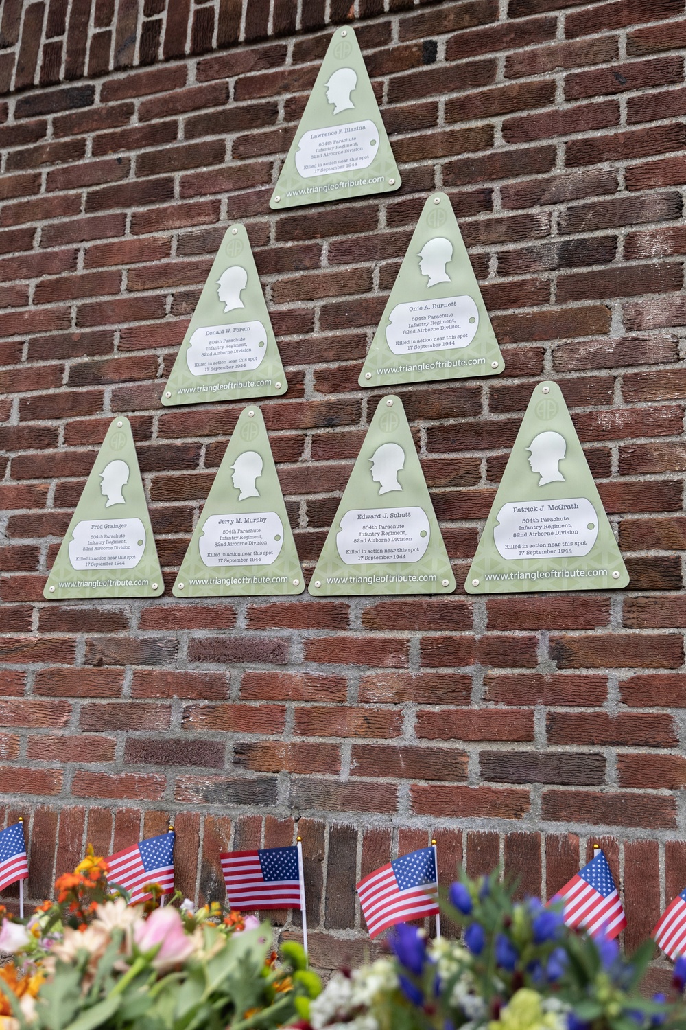 A brick wall displays seven green triangular memorial plaques from 'triangleoftribute.com', each bearing a silhouette profile and honoring American soldiers killed in action on 17 September 1944. The soldiers were members of the 504th Parachute Infantry Regiment, 82nd Airborne Division. Small American flags and flowers have been placed at the base of the wall as a tribute.