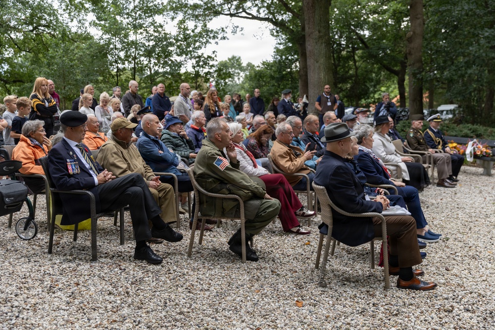 An outdoor memorial or commemorative ceremony is taking place in a wooded setting, with a large crowd of attendees seated on chairs arranged on a gravel surface. Several individuals in the front rows are wearing military uniforms, medals, and period attire, suggesting a WWII remembrance event. Dignitaries in formal military dress uniforms are visible on the right side, and floral arrangements can be seen in the background.