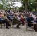 An outdoor memorial or commemorative ceremony is taking place in a wooded setting, with a large crowd of attendees seated on chairs arranged on a gravel surface. Several individuals in the front rows are wearing military uniforms, medals, and period attire, suggesting a WWII remembrance event. Dignitaries in formal military dress uniforms are visible on the right side, and floral arrangements can be seen in the background.