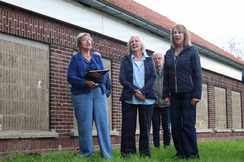 Four people are standing outdoors in front of a long, single-story brick building with boarded or paneled windows. Three women in the foreground appear to be speaking or singing, with one holding a tablet or folder. The setting suggests a group visit, tour, or outdoor gathering at what appears to be an older or historic structure.