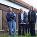 Four people are standing outdoors in front of a long, single-story brick building with boarded or paneled windows. Three women in the foreground appear to be speaking or singing, with one holding a tablet or folder. The setting suggests a group visit, tour, or outdoor gathering at what appears to be an older or historic structure.