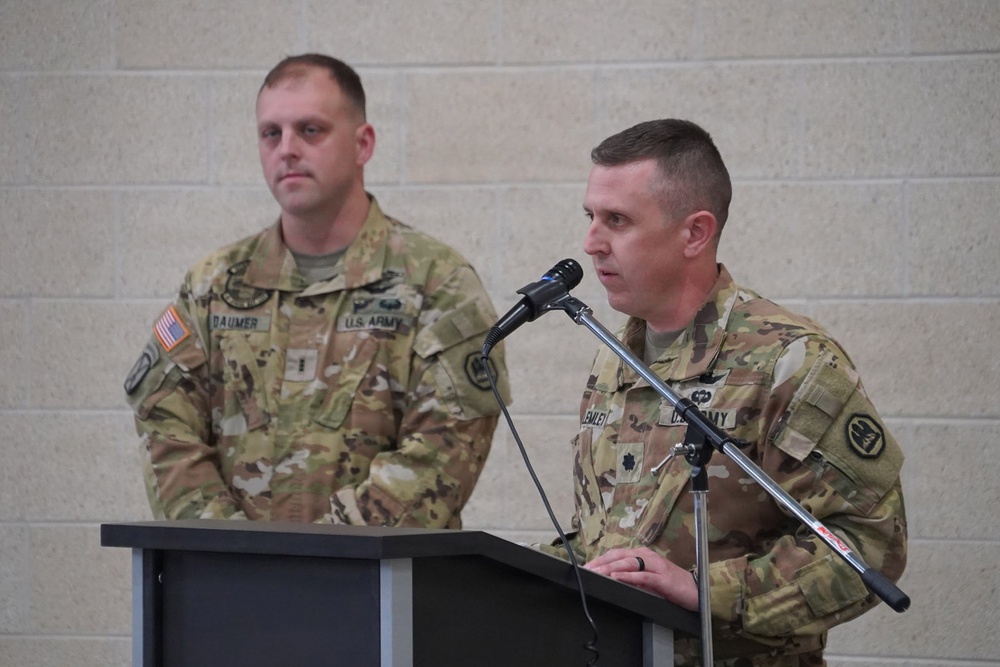 Two U.S. Army soldiers in OCP (Operational Camouflage Pattern) uniforms are participating in a formal military ceremony or briefing. One soldier is speaking at a podium with a microphone, while the other stands attentively in the background. Name tapes visible on their uniforms read 'DAUMER' and partially visible on the speaker.